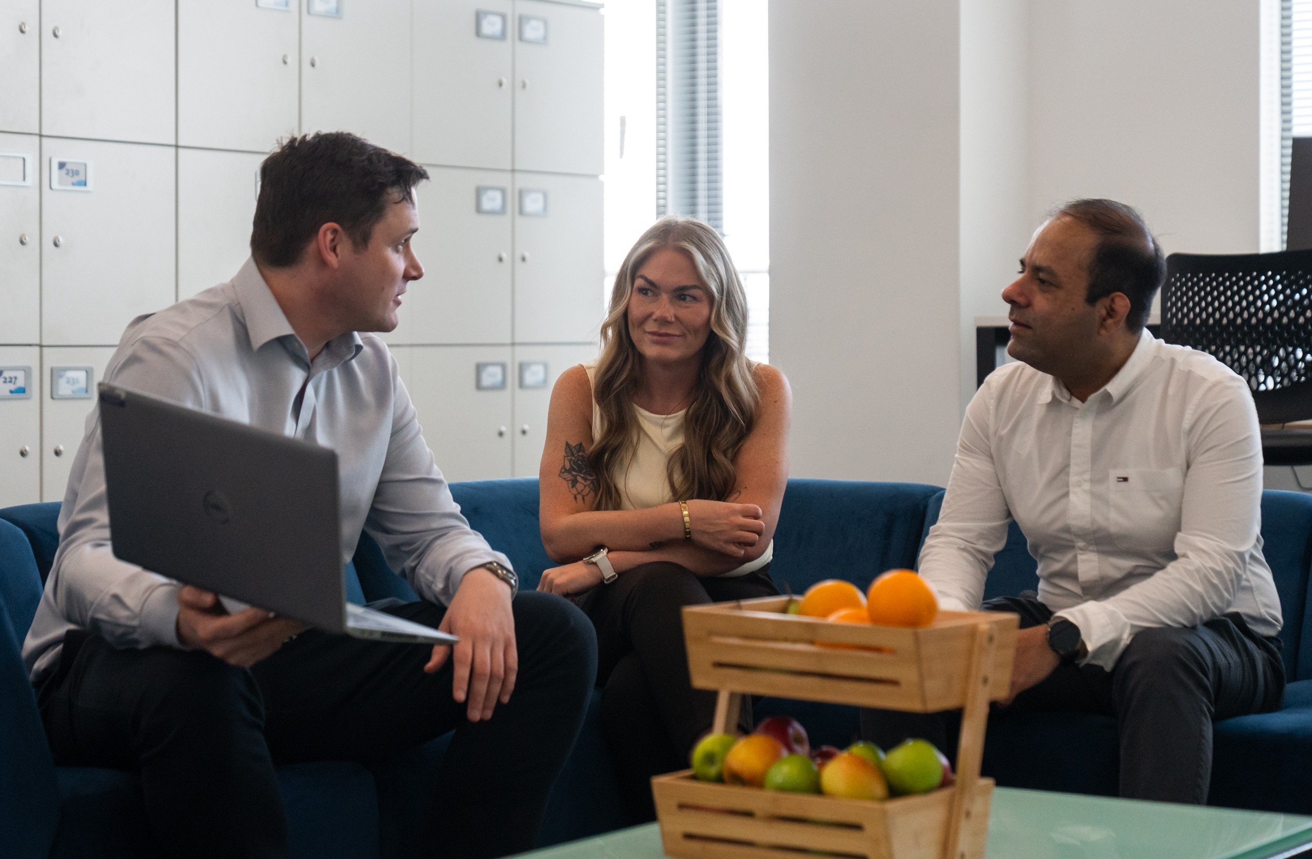 A woman and two men sitting talking in a breakout area with a basket of fruit in the foreground and lockers in the background