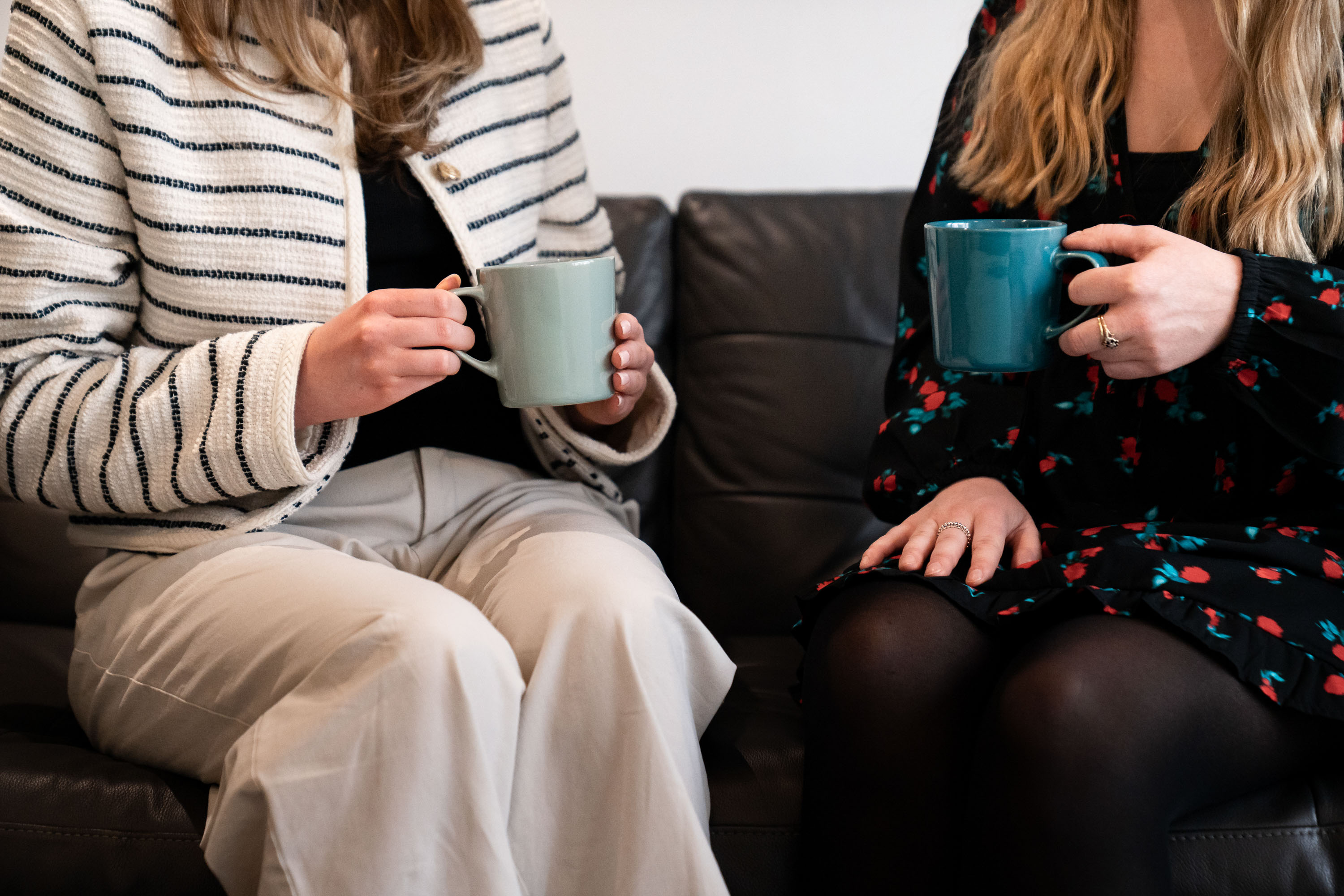 Close-up of two women, faces not visible, sitting on a sofa, each holding a mug
