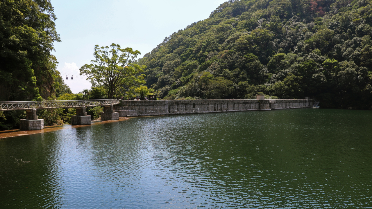 A calm reservoir bordered by a stone dam and a narrow bridge, surrounded by forested hills under a clear sky