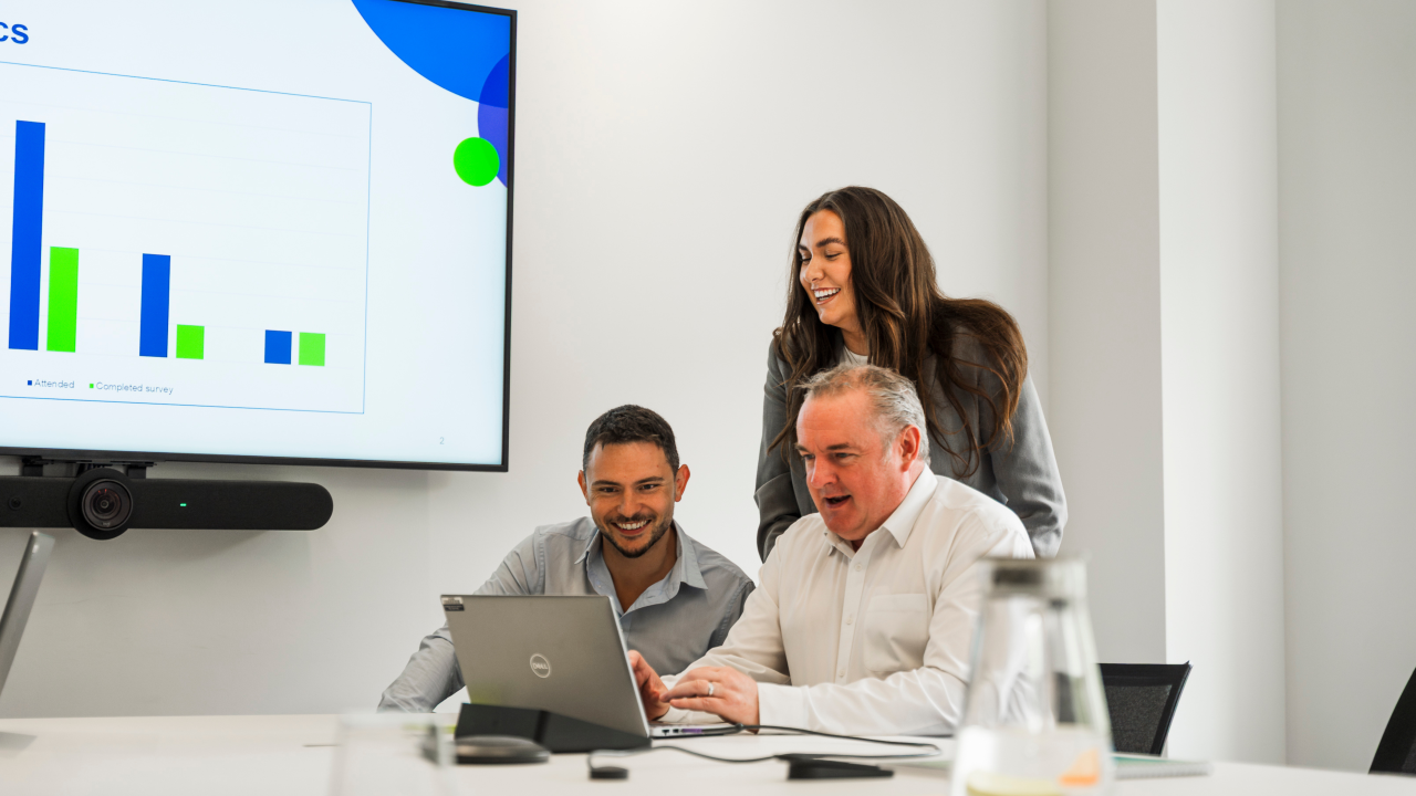 Three colleagues reviewing data on a laptop in a meeting room with a chart displayed on a screen
