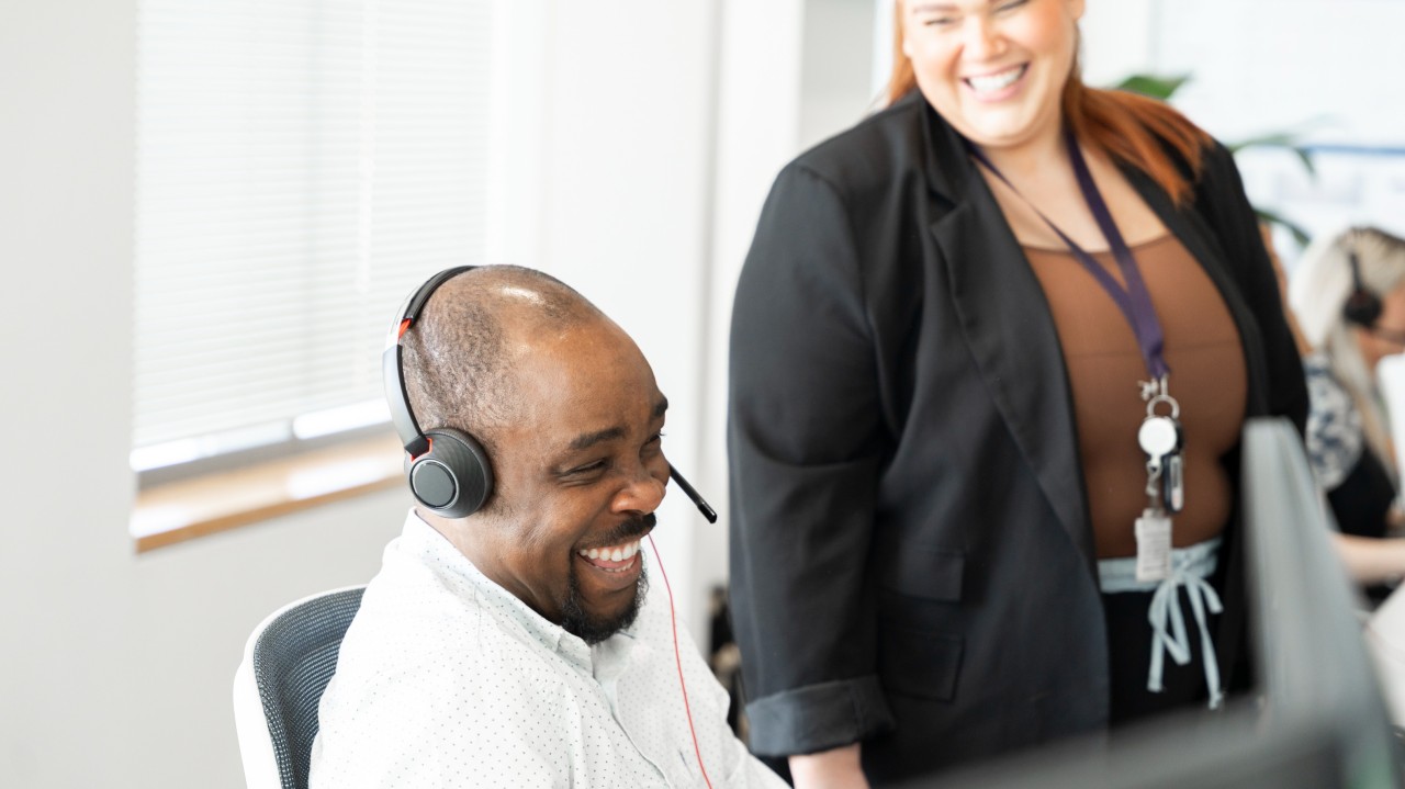 Two smiling colleagues in an office setting: one seated at a desk wearing a headset and the other standing beside them.
