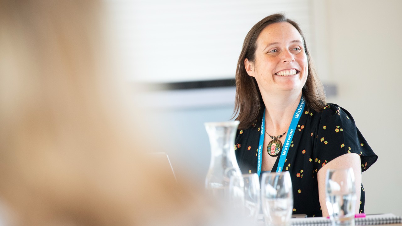 Jo wearing a black shirt with a colourful pattern and a blue lanyard sitting at a table with several empty glasses and a glass pitcher, smiling.