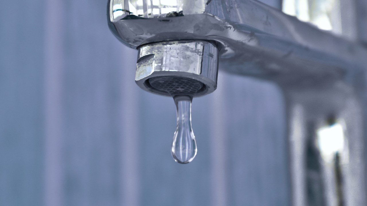 Close-up of a chrome tap with a single droplet of water hanging from the spout, set against a softly blurred blue background.