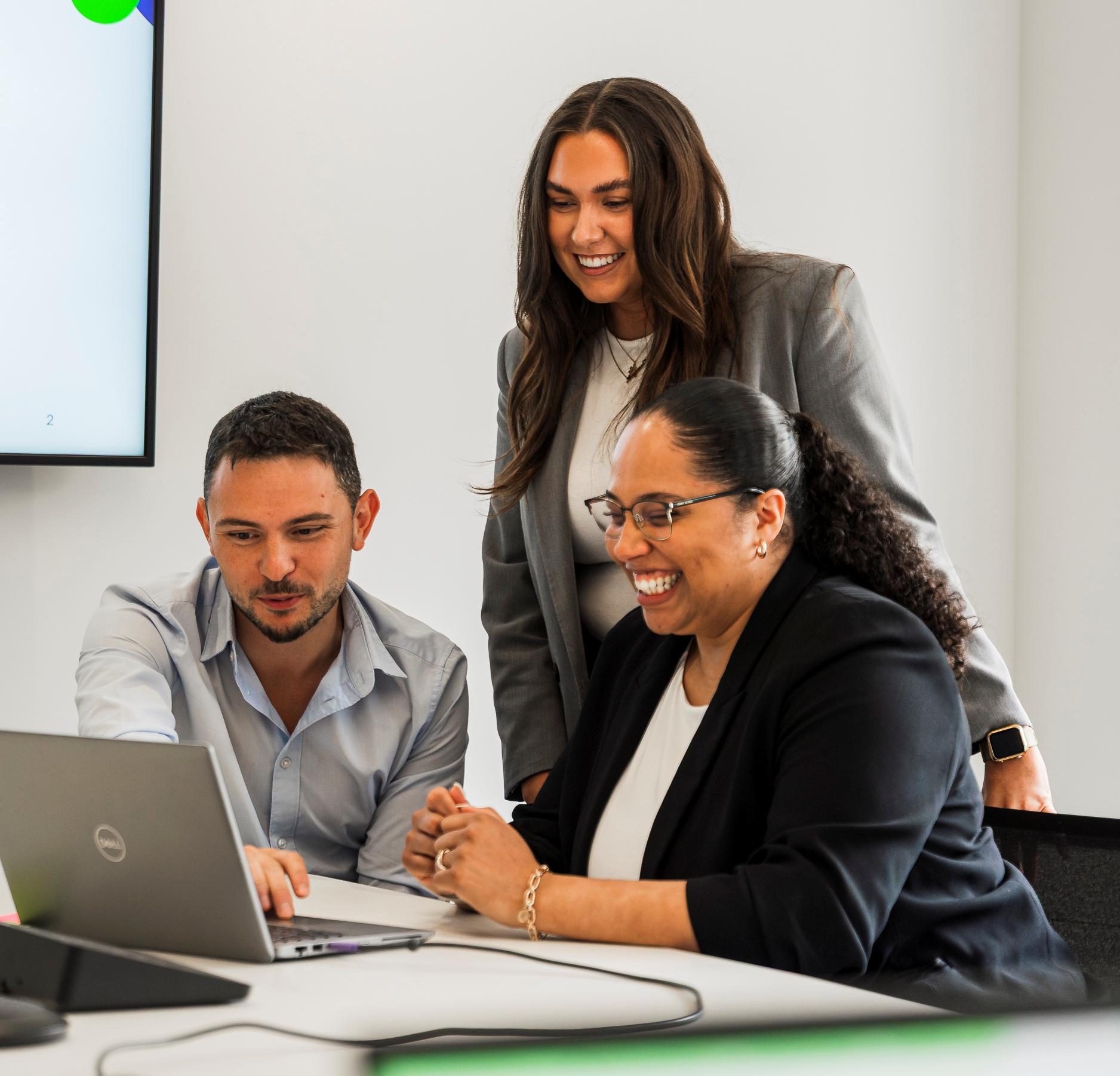 One woman standing and a man and woman seated looking at a laptop screen and smiling