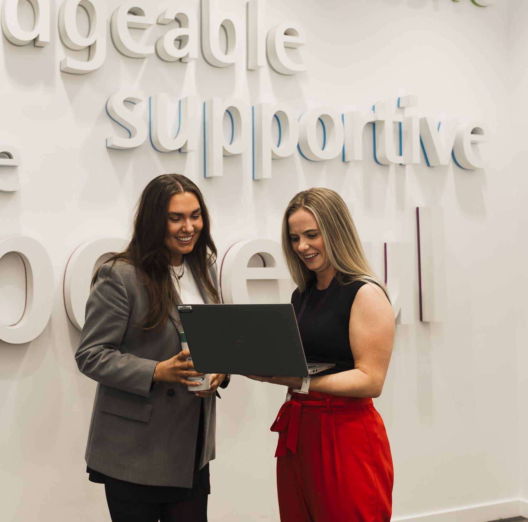 Two smiling women standing in front of a sign with the word "supportive" visible while looking at a laptop