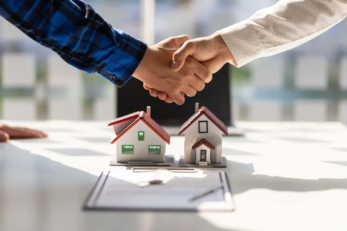 Close-up of two people shaking hands above paperwork and two small models of homes