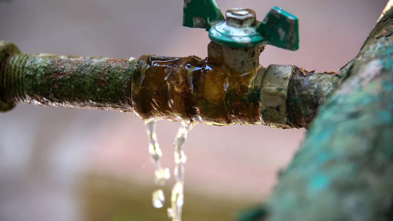 A close-up of a corroded outdoor water valve leaking a steady stream of water from a crack in the pipe, with droplets running down