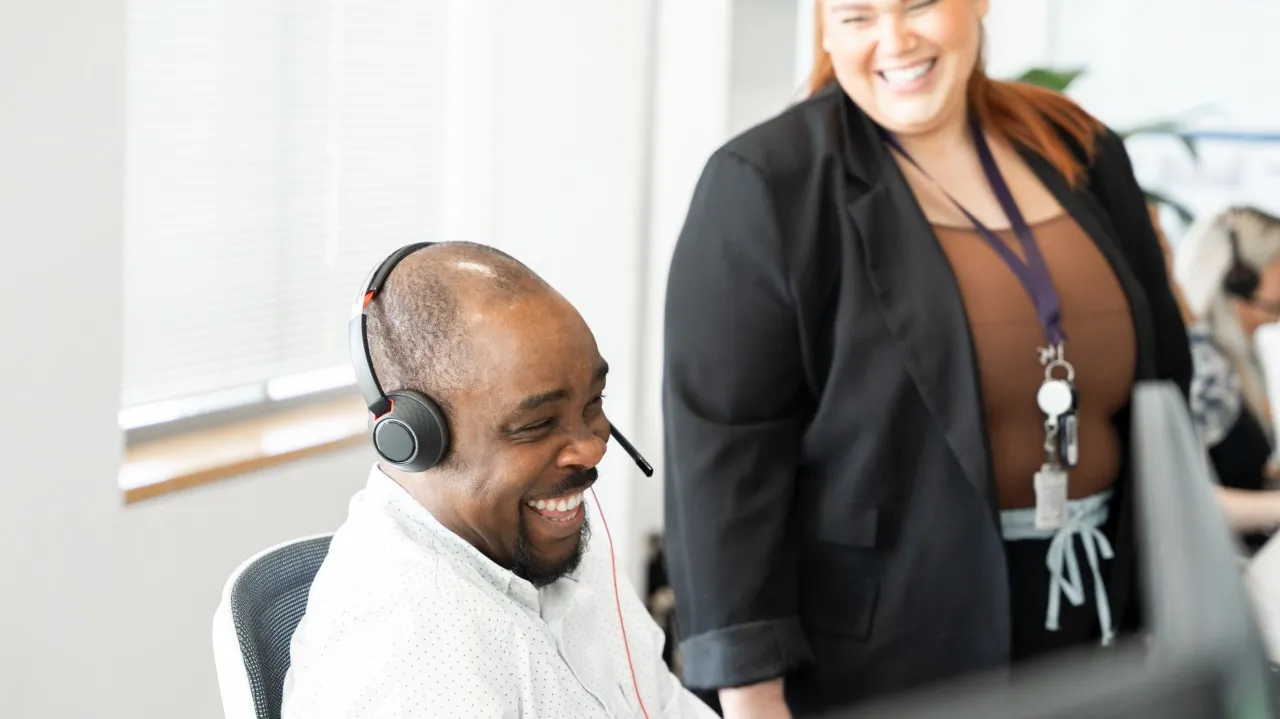 Two smiling colleagues in an office setting: one seated at a desk wearing a headset and the other standing beside them.