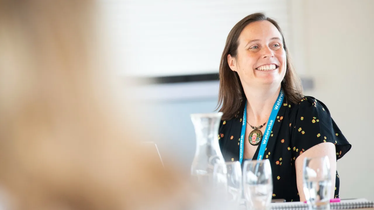 Jo wearing a black shirt with a colourful pattern and a blue lanyard sitting at a table with several empty glasses and a glass pitcher, smiling.