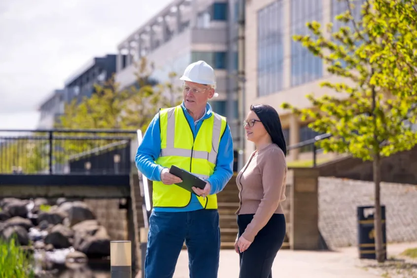 Man in hard hat and hi-vis jacket speaking to woman outside with buildings and landscaping in background