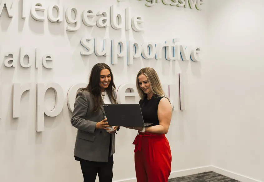 Two smiling women standing in front of a sign with the word "supportive" visible while looking at a laptop