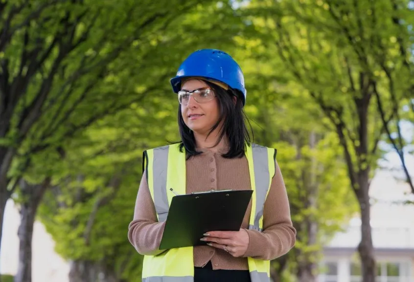 Woman in blue hard hat holding clipboard with green trees in background
