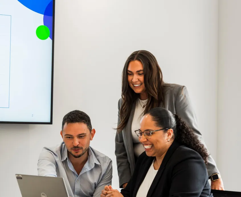 One woman standing and a man and woman seated looking at a laptop screen and smiling