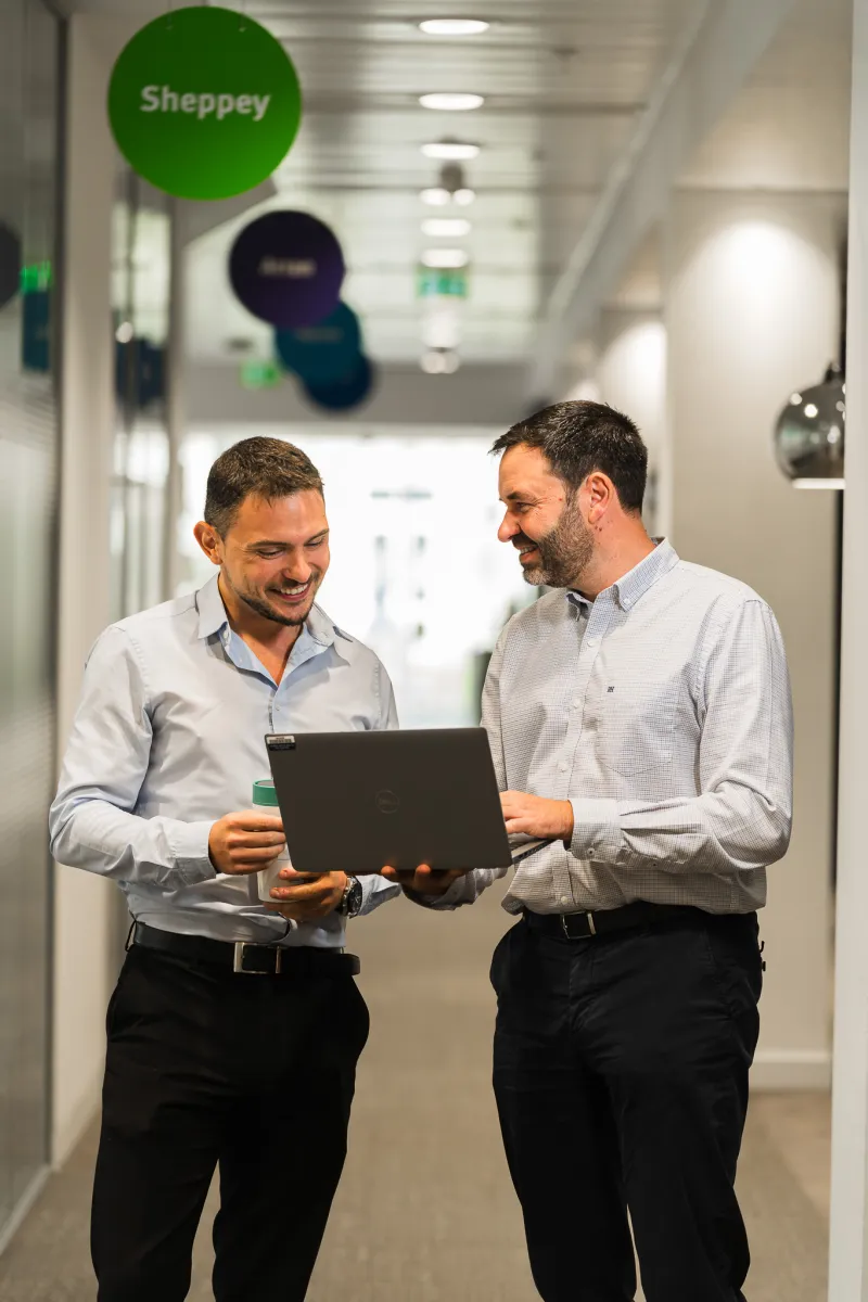 Two smiling men standing in an office space looking at a laptop