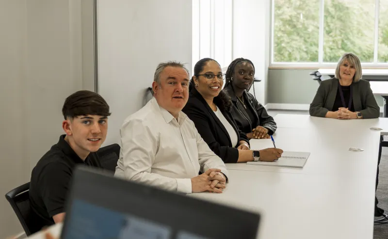 Three women and two men seated around a large conference table listening to someone off screen
