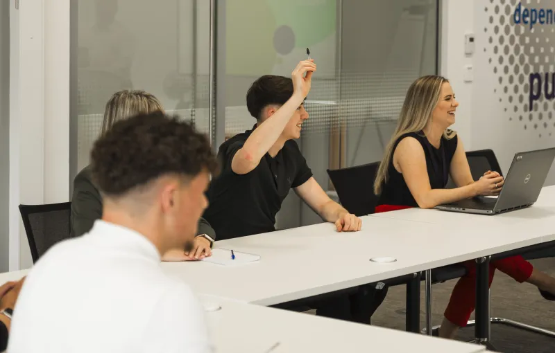 Two women, one of which obscured, and two young men, one with his hand raised, seated around a conference table.