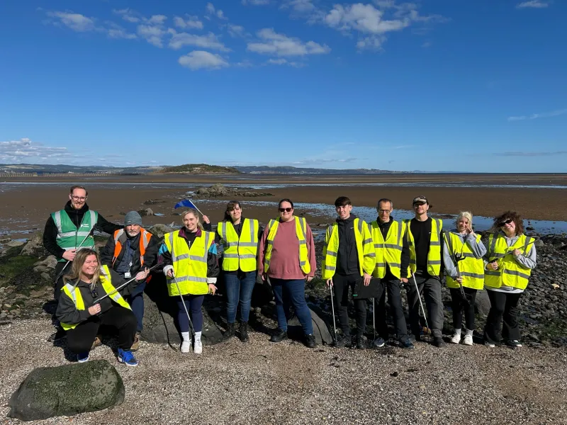 A group of 11 smiling people on a beach, wearing hi-vis vests and holding litter pickers