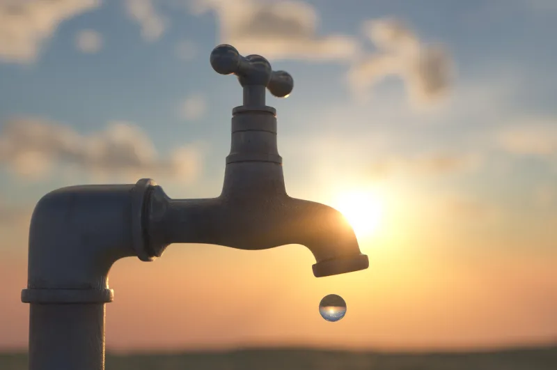 Single round drop of water falling from a dry tap outdoors with low sun in blue sky directly behind