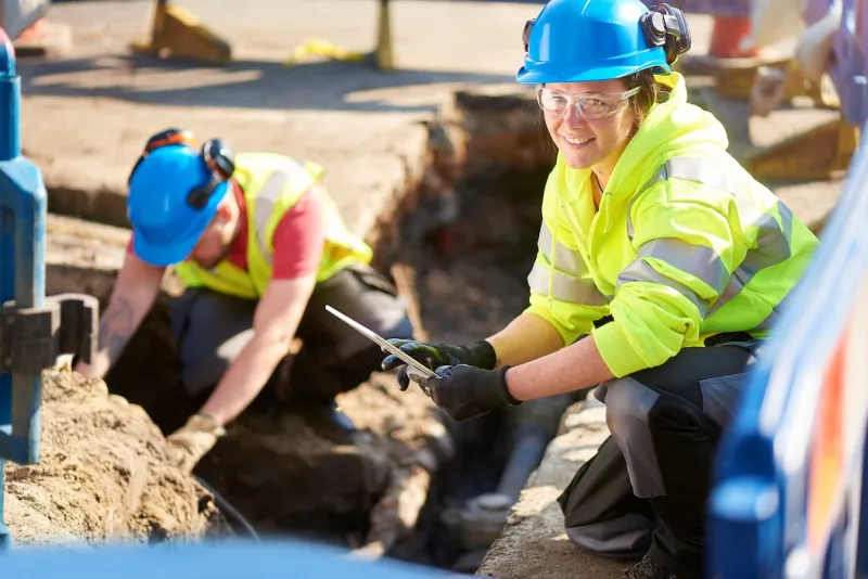 Female engineer wearing a blue hard hat and crouching over a trench on a construction site.
