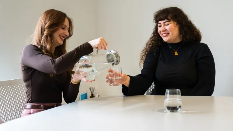Two people at a table pouring drinking water from a glass jug into a tumbler