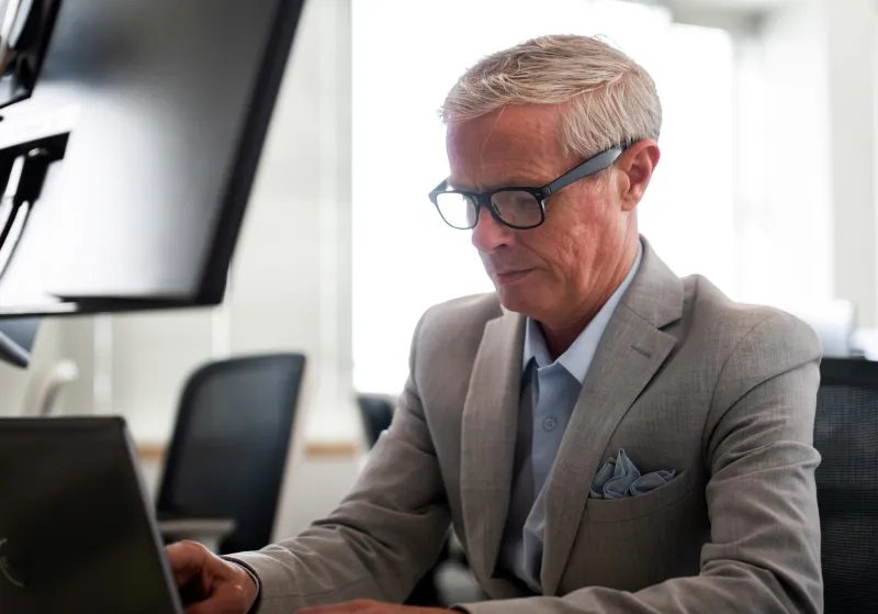 Man in suit with no tie working at laptop with large monitor above