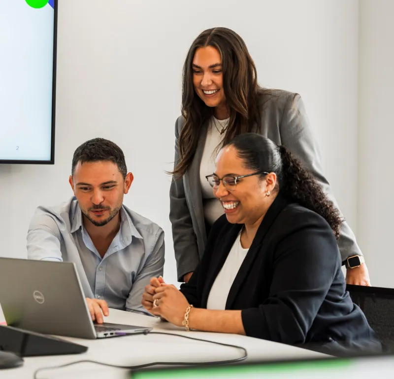 One woman standing and a man and woman seated looking at a laptop screen and smiling