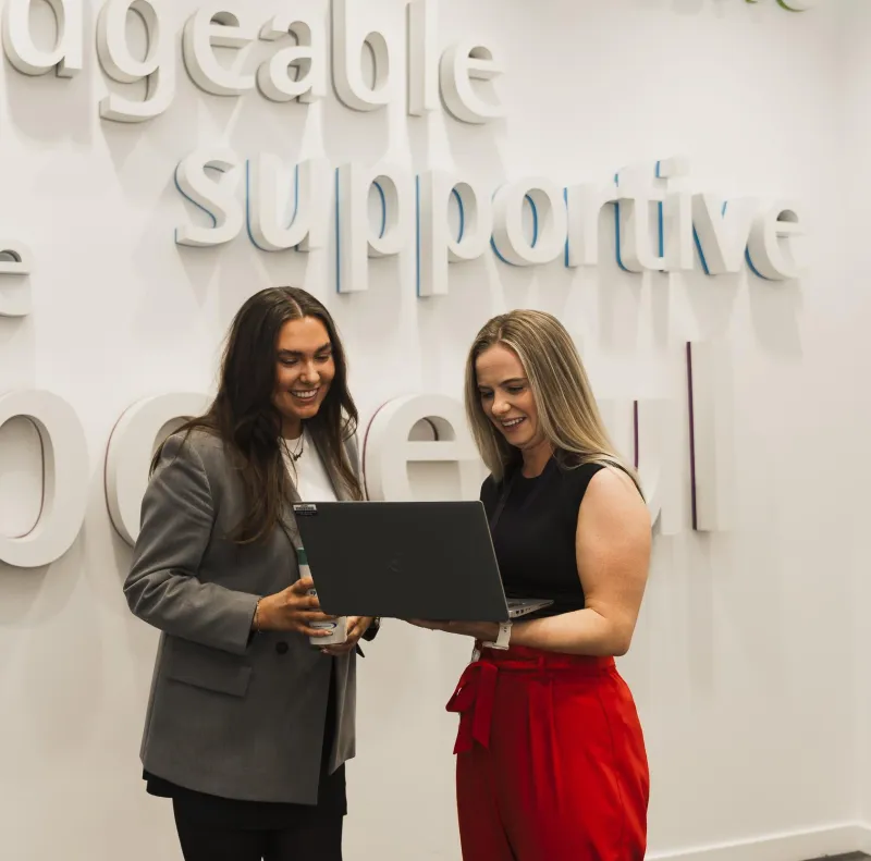 Two smiling women standing in front of a sign with the word "supportive" visible while looking at a laptop
